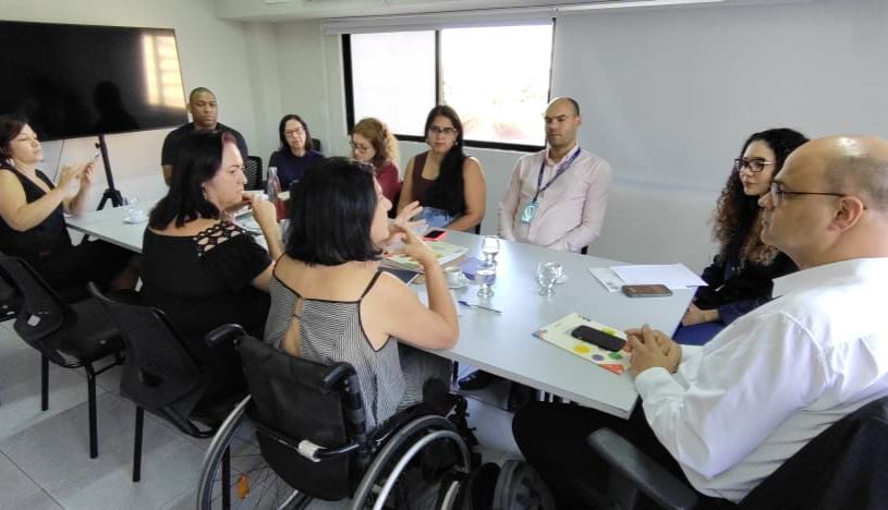 Grupo em reunião, com intérprete de libras, mulher em cadeira de rodas e clima colaborativo.