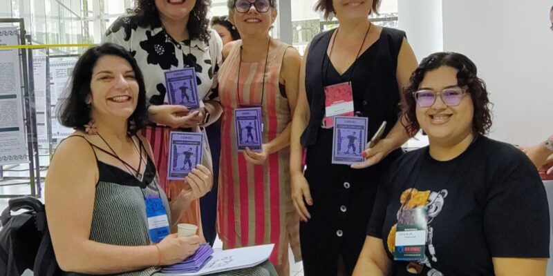Cinco mulheres sorrindo, segurando cordéis roxos com desenho de cadeirante e a palavra CORDEL.