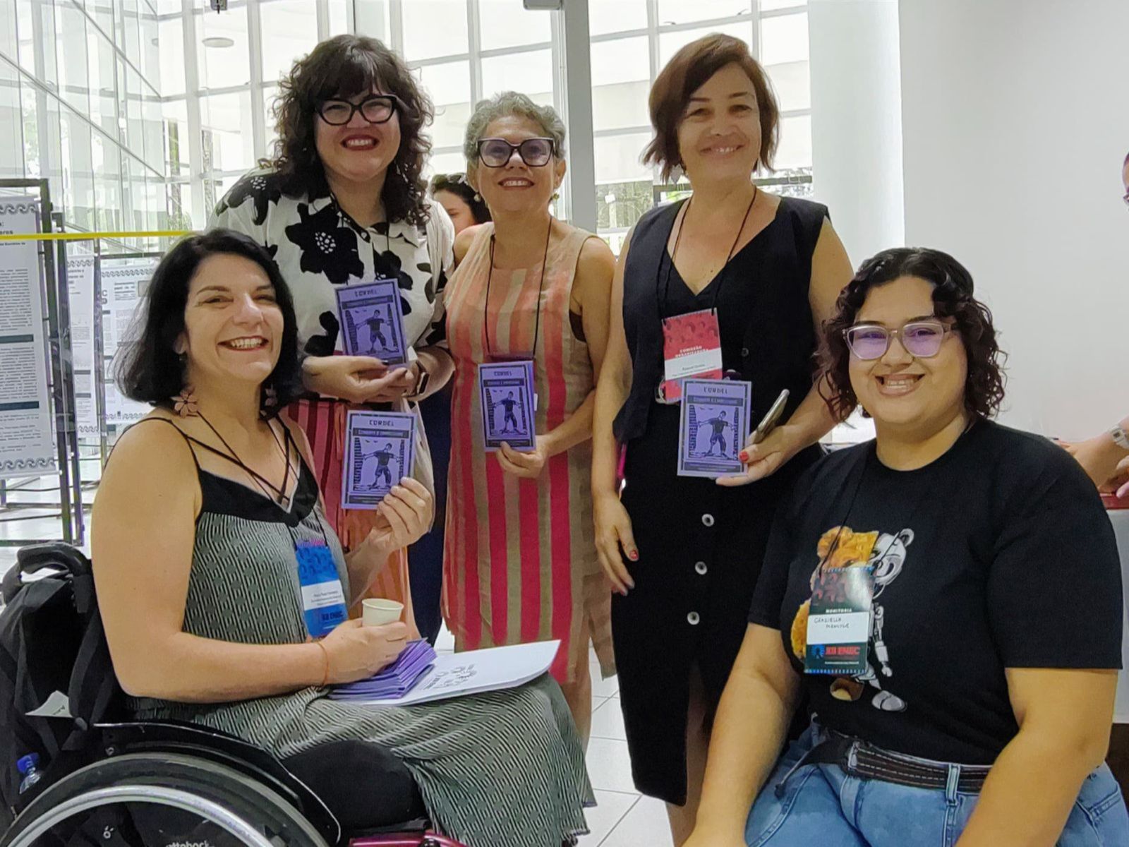 Cinco mulheres sorrindo, segurando cordéis roxos com desenho de cadeirante e a palavra CORDEL.