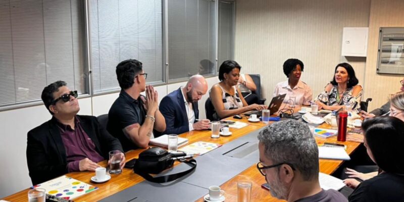 Reunião com doze pessoas em mesa de escritório, papéis, notebooks, café e ambiente colaborativo.
