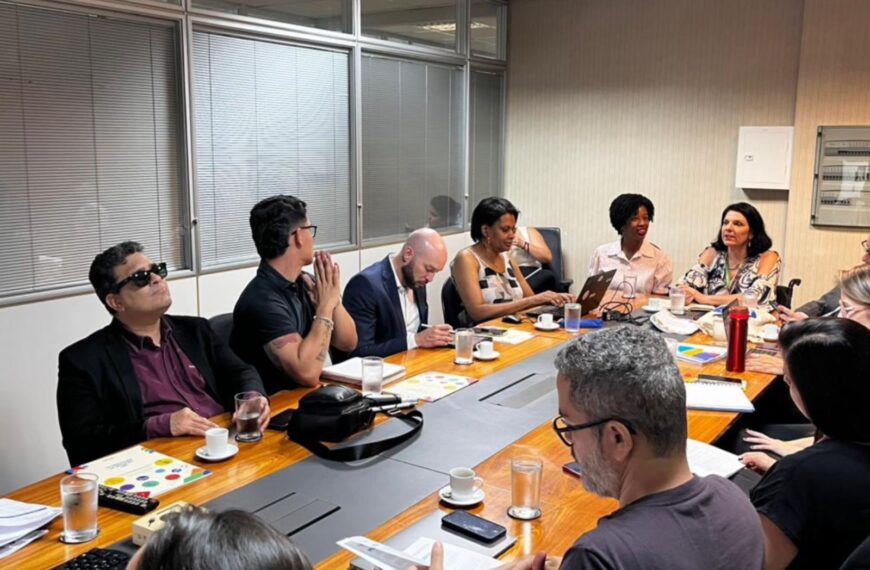 Reunião com doze pessoas em mesa de escritório, papéis, notebooks, café e ambiente colaborativo.