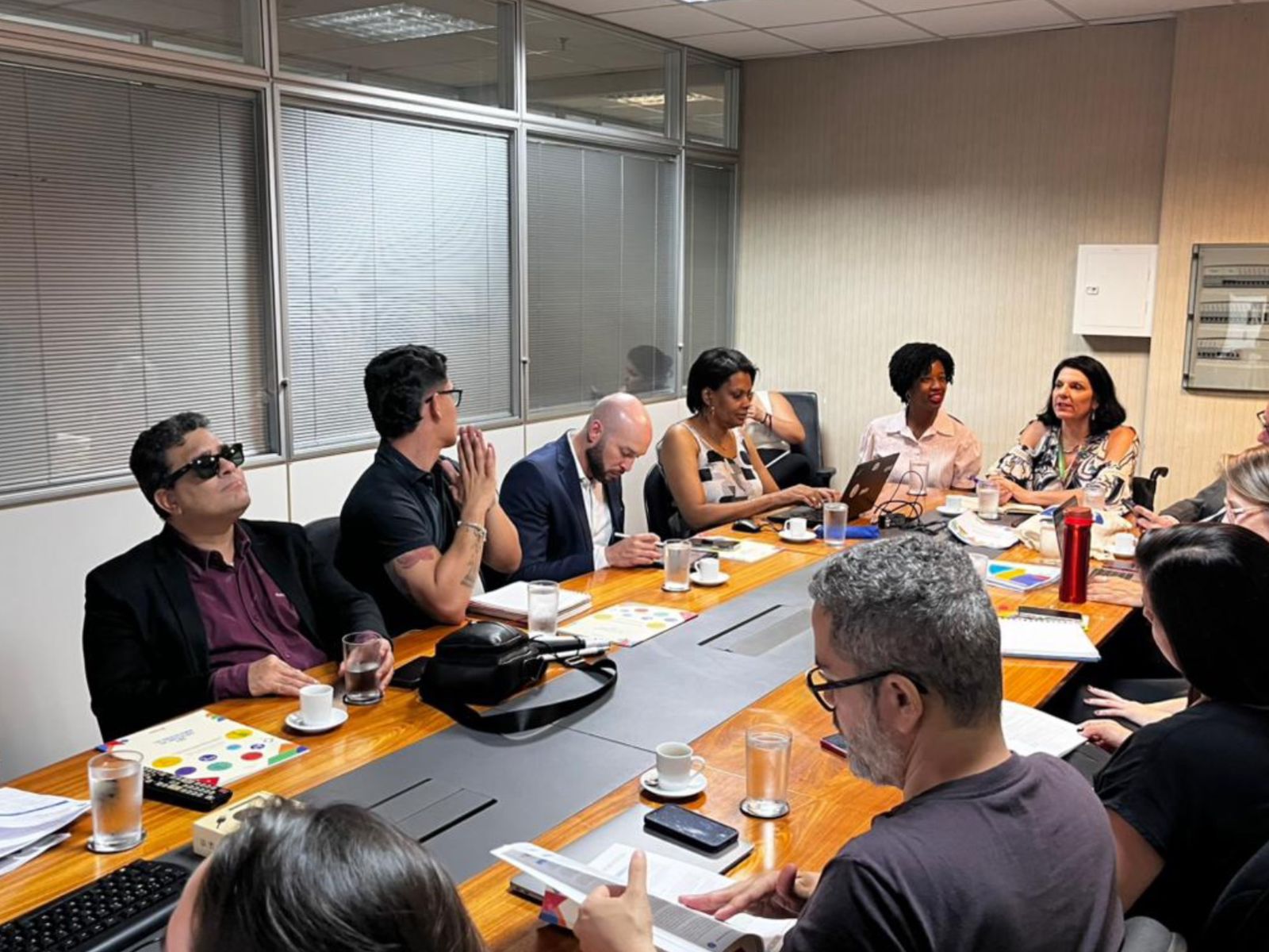 Reunião com doze pessoas em mesa de escritório, papéis, notebooks, café e ambiente colaborativo.