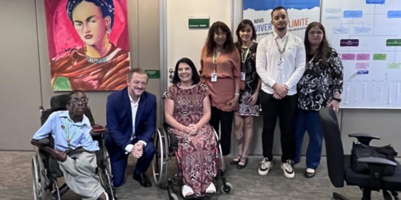 Sete pessoas em grupo, incluindo duas em cadeiras de rodas, em escritório com pôster da Frida.