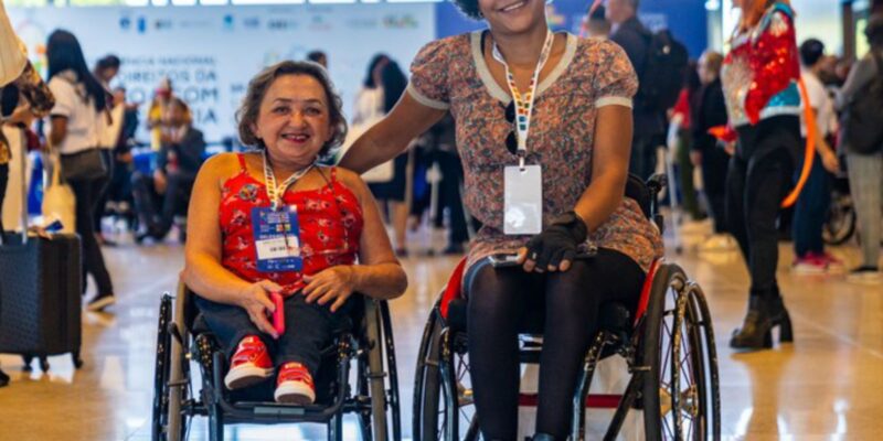 Duas mulheres em cadeiras de rodas sorrindo, com crachá no peito, em um evento movimentado.