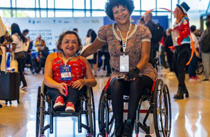 Duas mulheres em cadeiras de rodas sorrindo, com crachá no peito, em um evento movimentado.