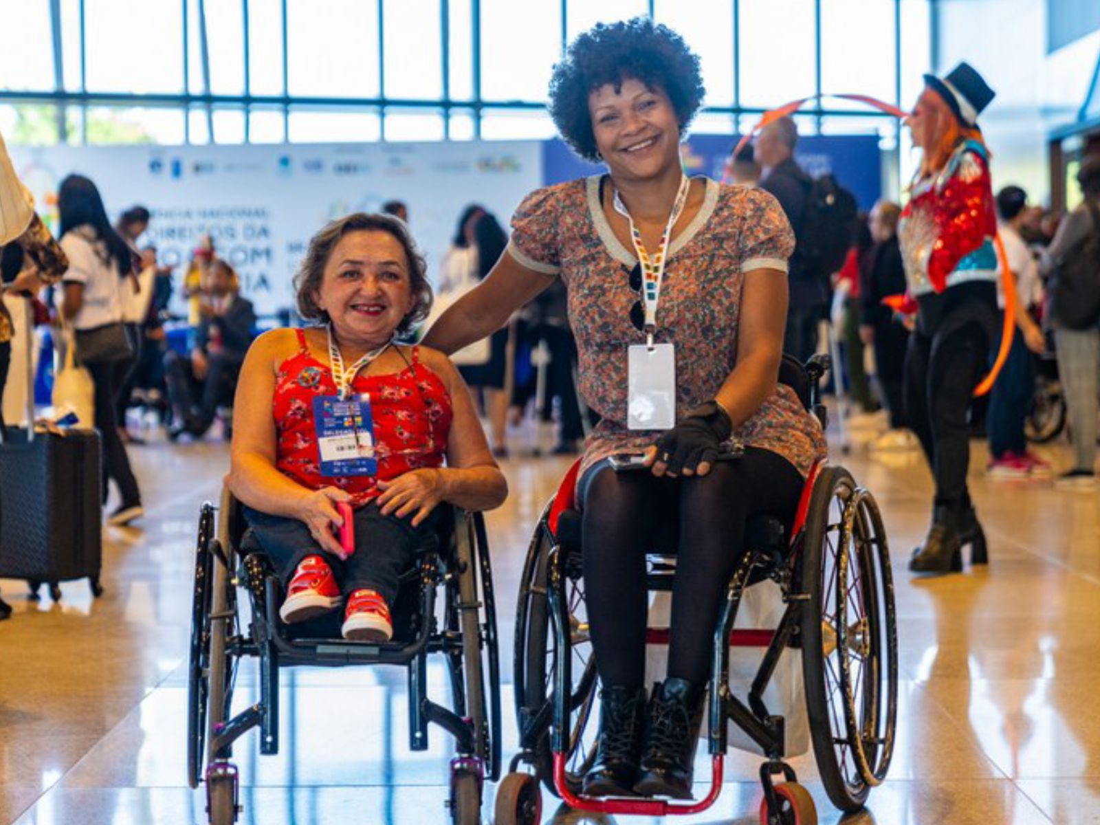 Duas mulheres em cadeiras de rodas sorrindo, com crachá no peito, em um evento movimentado.