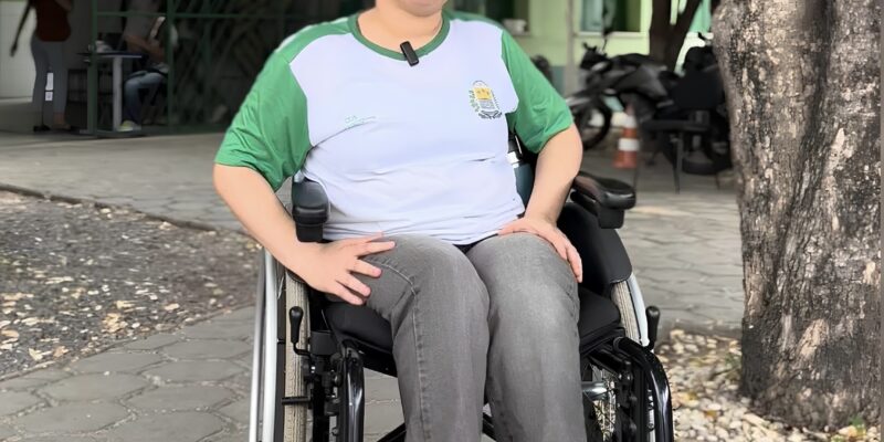 Mulher de cabelo curto em cadeira de rodas, usando camiseta escolar, sentada sob árvore na calçada.