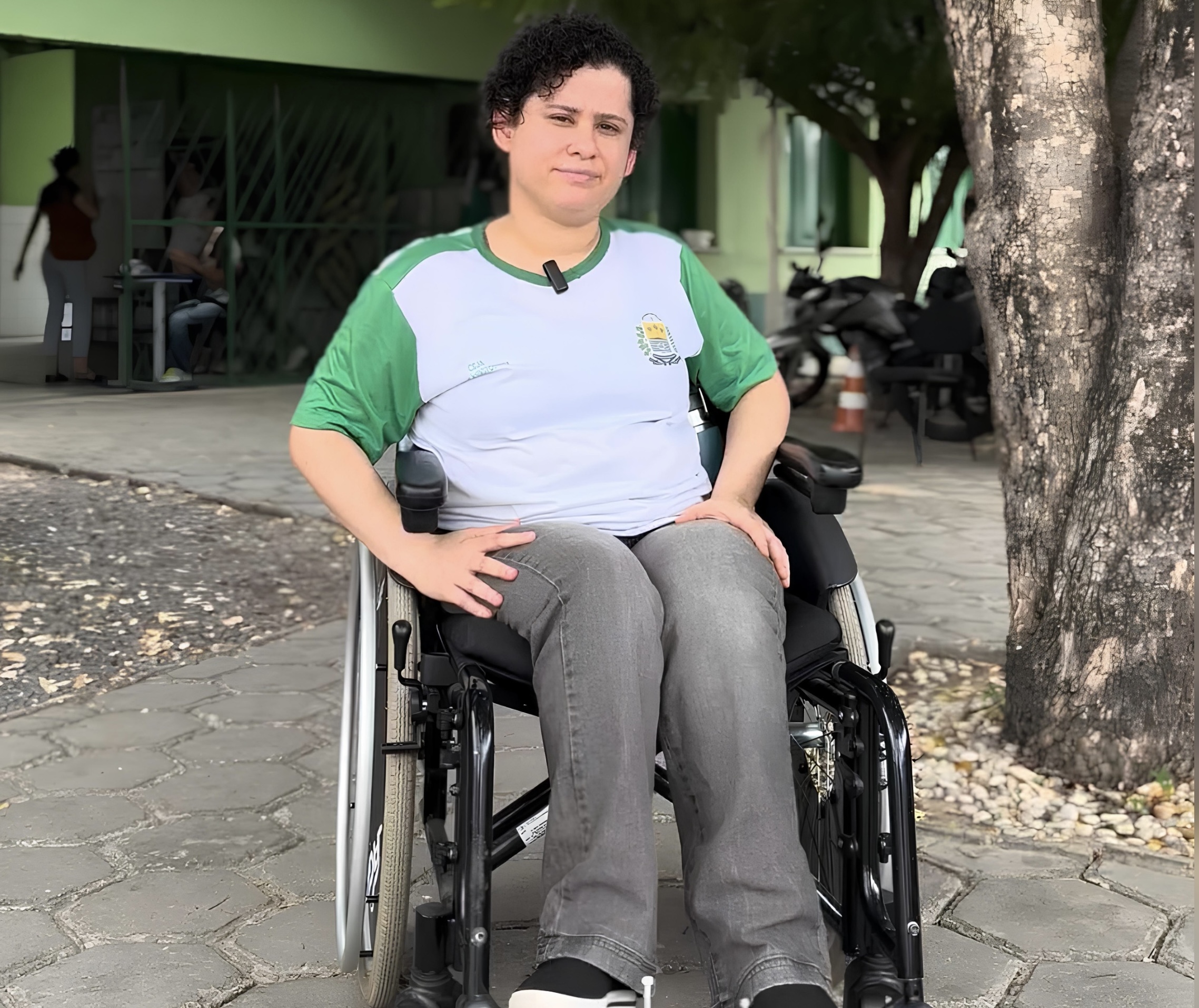 Mulher de cabelo curto em cadeira de rodas, usando camiseta escolar, sentada sob árvore na calçada.