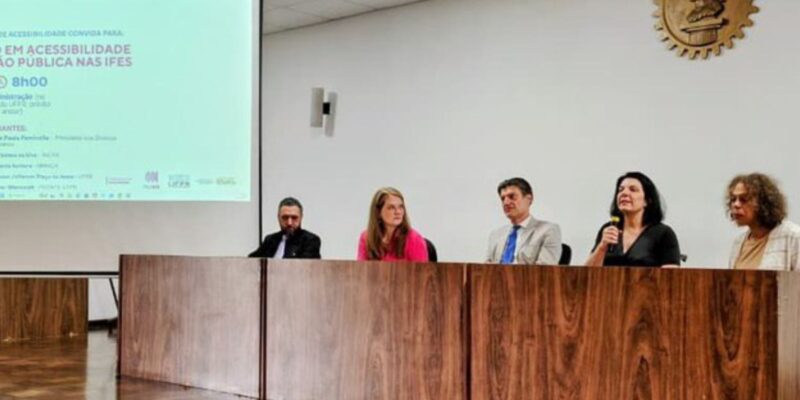 Cinco pessoas sentadas em mesa de cerimônia, uma fala ao microfone; tela exibe evento de acessibilidade.