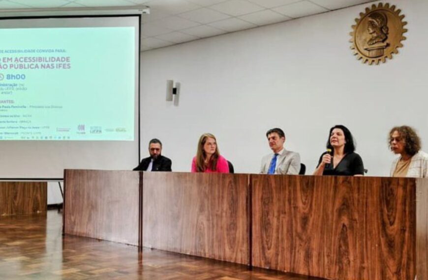 Cinco pessoas sentadas em mesa de cerimônia, uma fala ao microfone; tela exibe evento de acessibilidade.