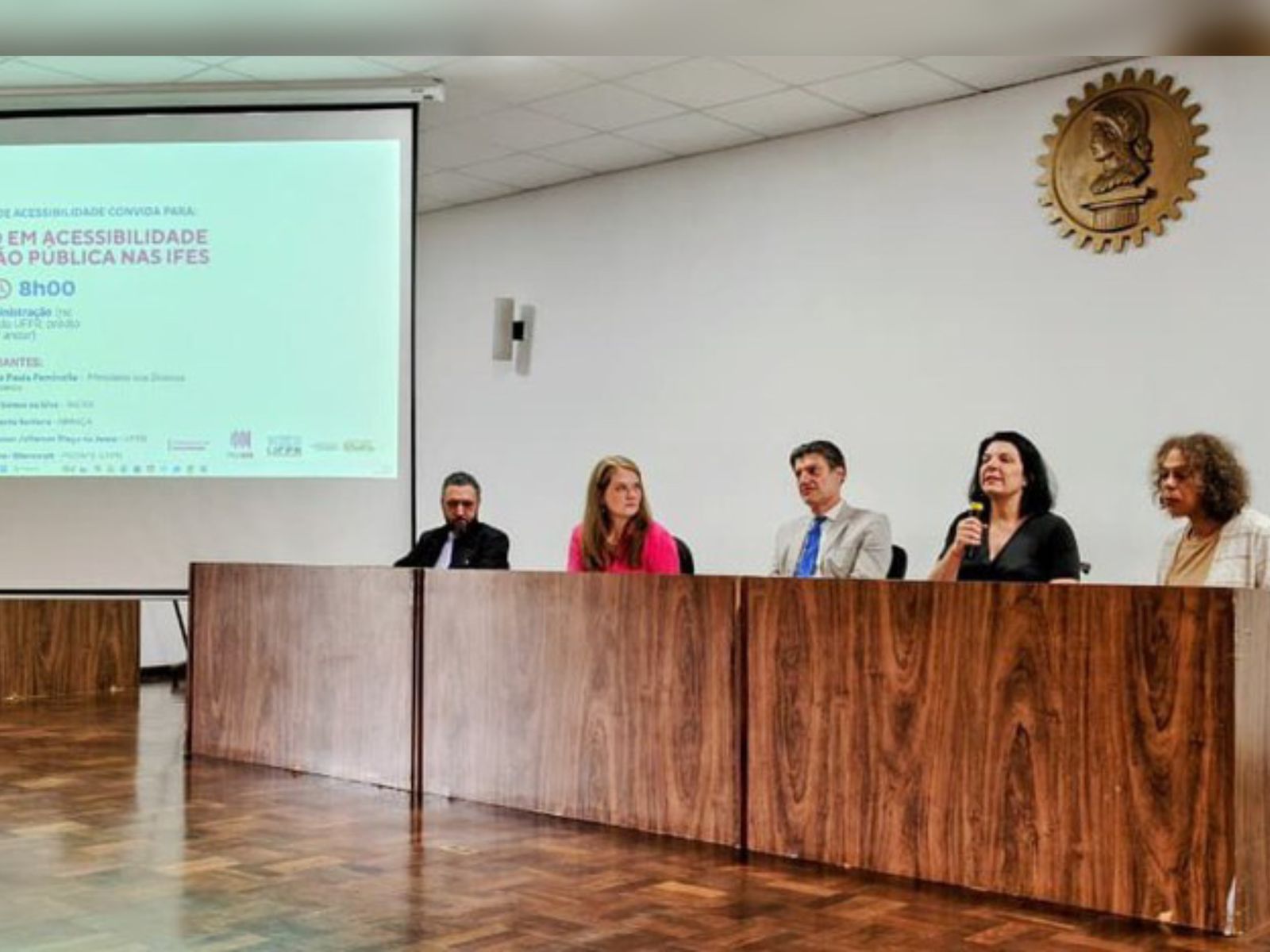 Cinco pessoas sentadas em mesa de cerimônia, uma fala ao microfone; tela exibe evento de acessibilidade.