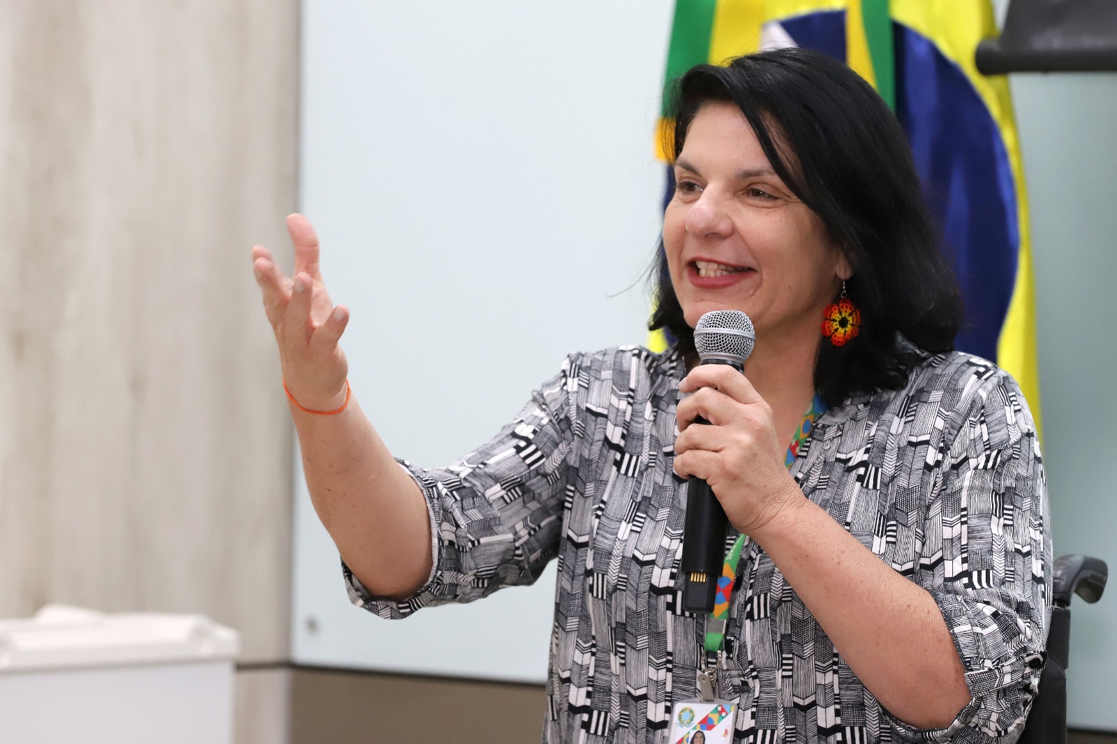 MDHC participa de lançamento da Rede Nacional de Pesquisa em Autismo e defende ciência para inclusão 3 A secretária Anna Paula, sorridente, fala ao microfone gesticulando, com bandeira do Brasil ao fundo.