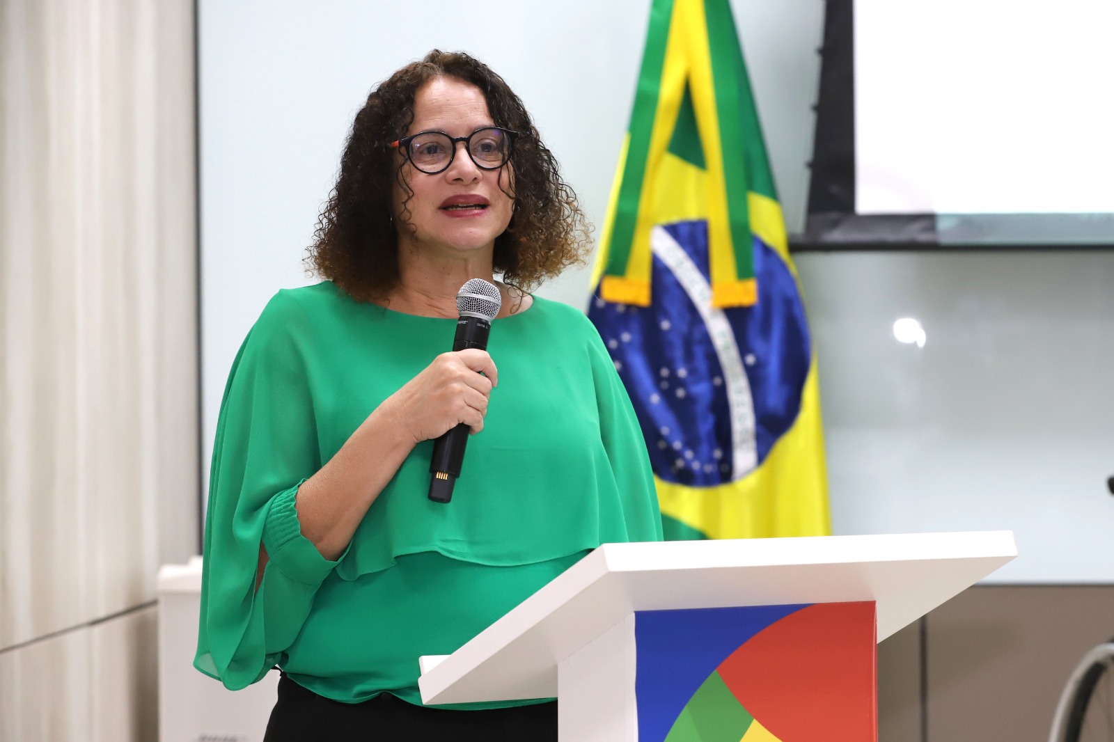 MDHC participa de lançamento da Rede Nacional de Pesquisa em Autismo e defende ciência para inclusão 5 Ministra Luciana Santos discursando em evento oficial, usando blusa verde, com bandeira do Brasil ao fundo.