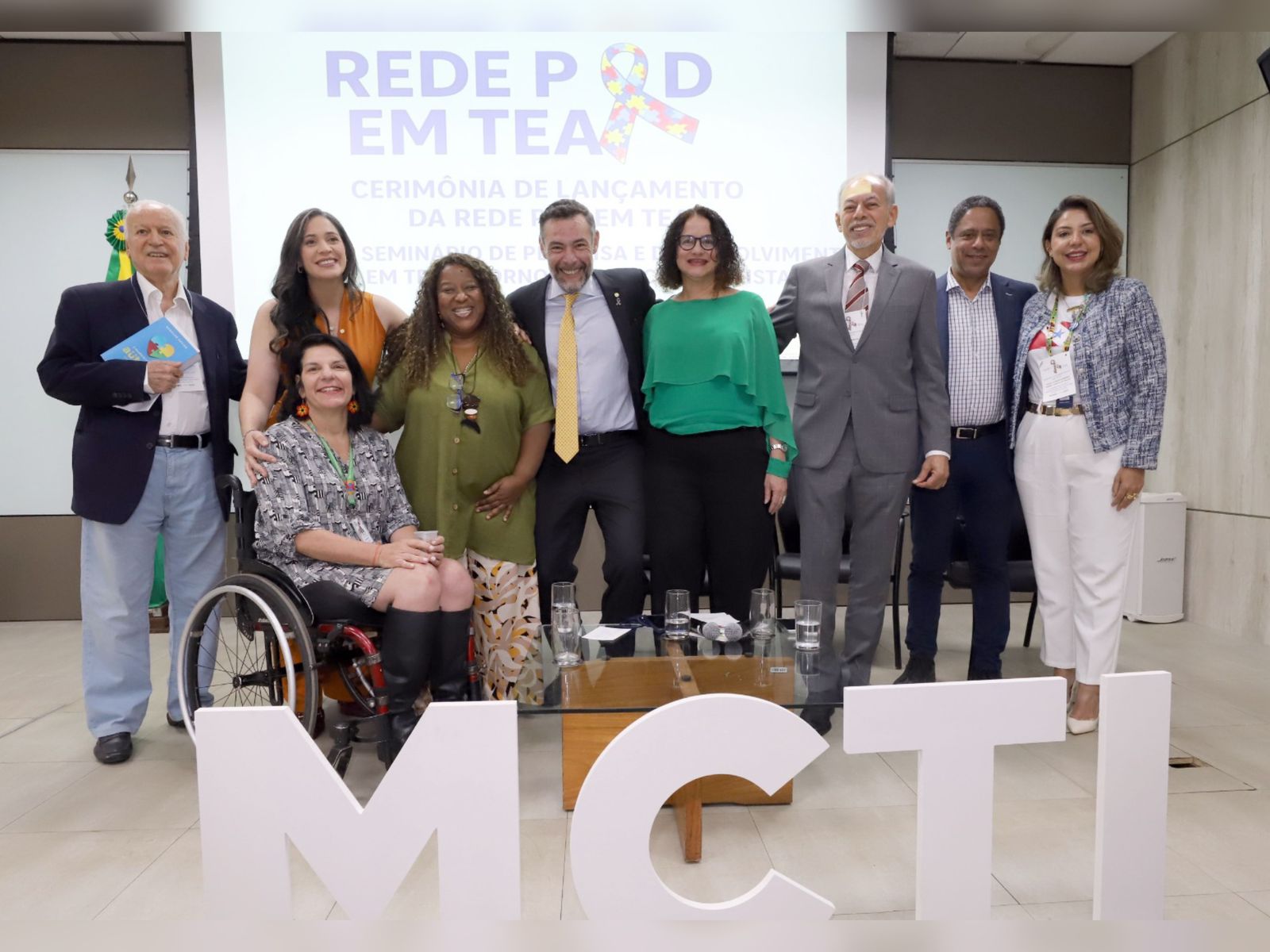Grupo de nove pessoas posa sorrindo em evento oficial sobre autismo, à frente das letras MCTI.