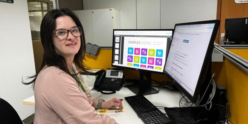 Mulher de óculos trabalha em mesa com dois monitores, teclado e telefone em escritório moderno.