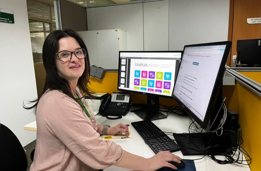 Mulher de óculos trabalha em mesa com dois monitores, teclado e telefone em escritório moderno.