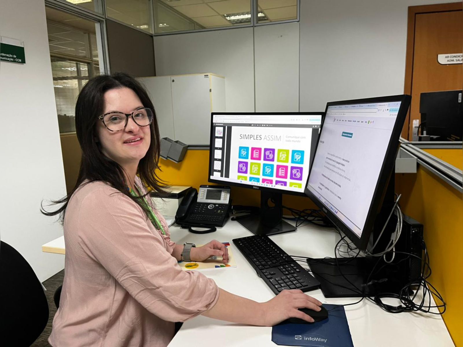 Mulher de óculos trabalha em mesa com dois monitores, teclado e telefone em escritório moderno.