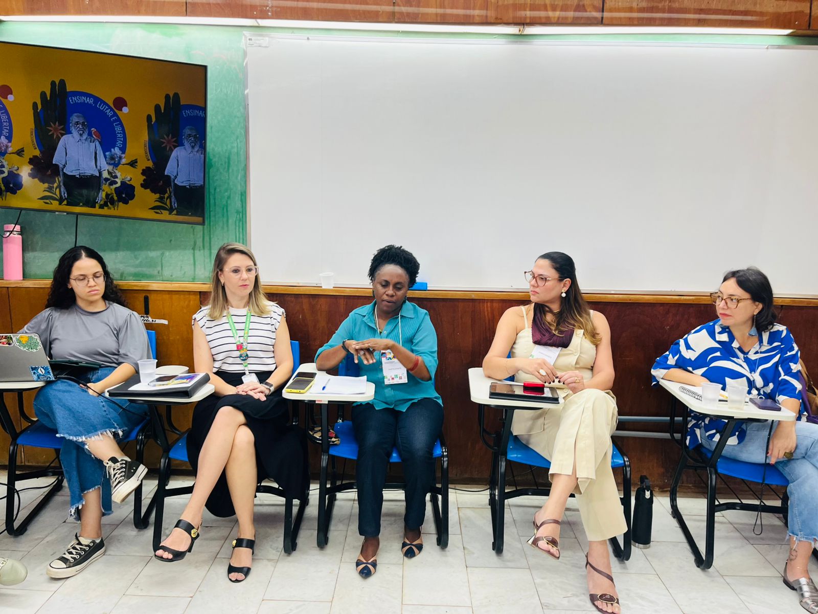 Cinco mulheres sentadas em carteiras escolares, conversando em uma sala com quadro branco atrás.