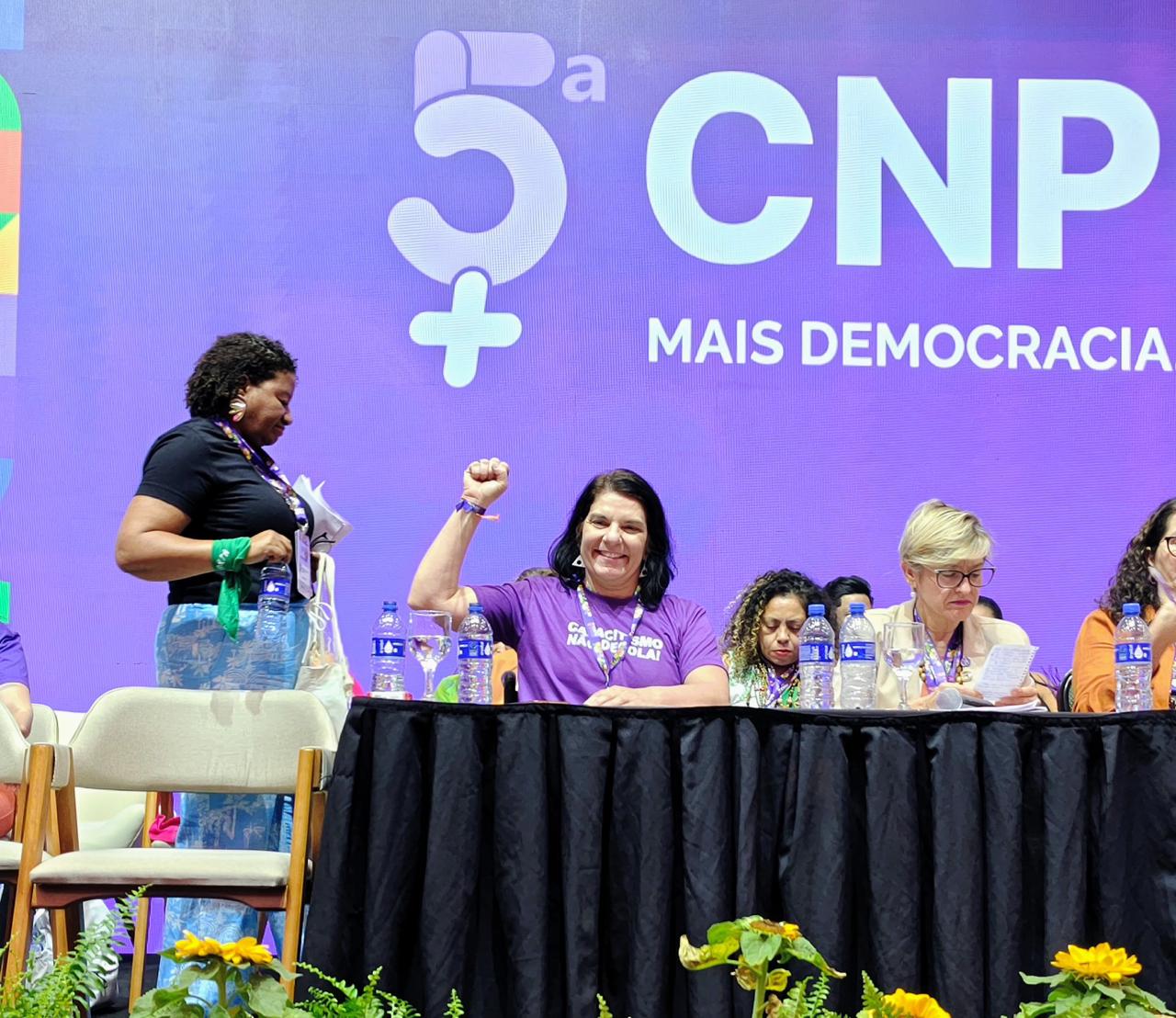 Mesa de mulheres em evento, fundo roxo, flores e frase “5ª CNP Mais Democracia” ao fundo.