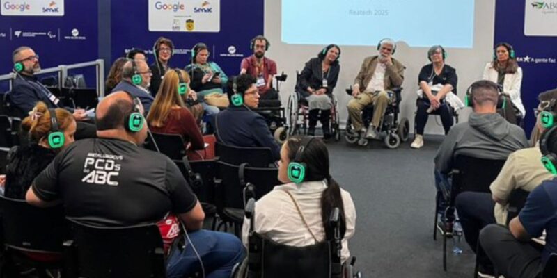 Roda de conversa inclusiva, pessoas com fones de ouvido, algumas em cadeiras de rodas, painel ao fundo.