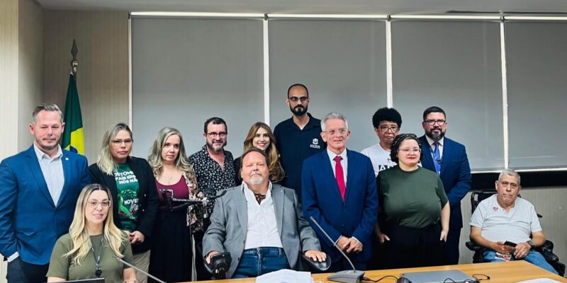 Quinze pessoas reunidas em sala de reunião, incluindo duas em cadeiras de rodas, clima formal.