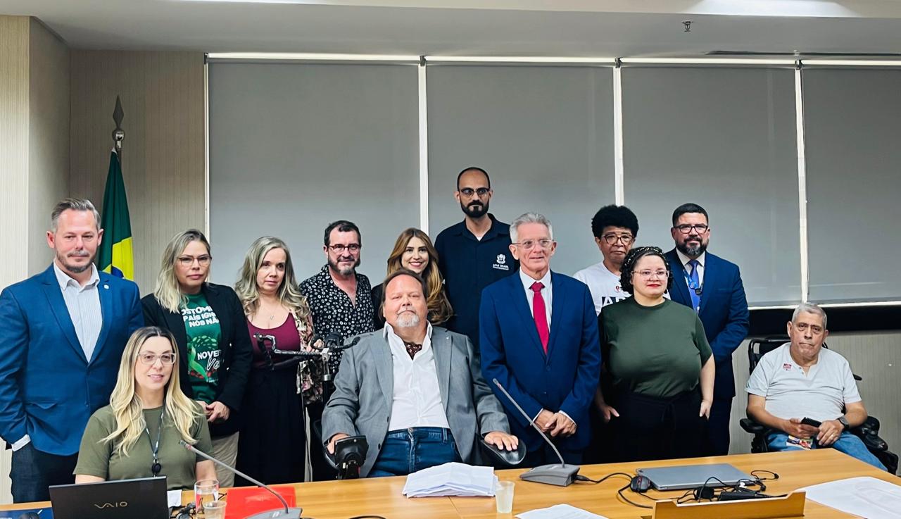 Quinze pessoas reunidas em sala de reunião, incluindo duas em cadeiras de rodas, clima formal.