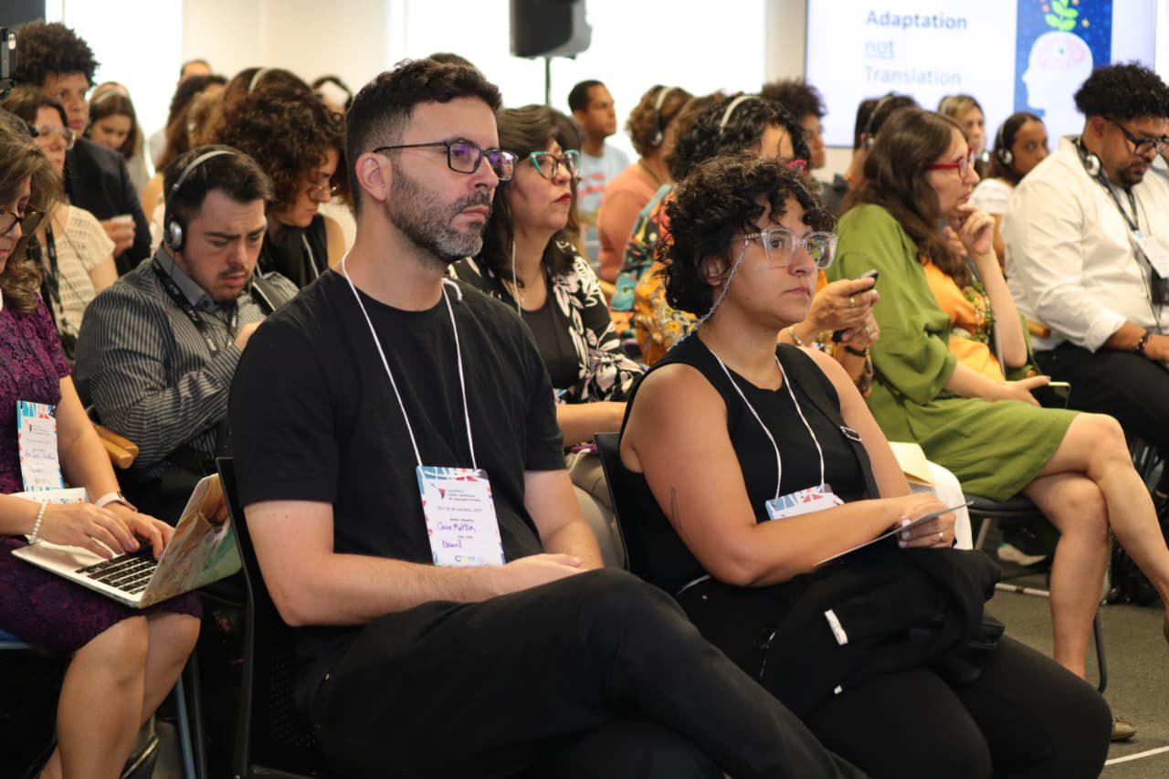 Pessoas sentadas em um evento, ouvindo atentas, algumas com fones e crachás pendurados.