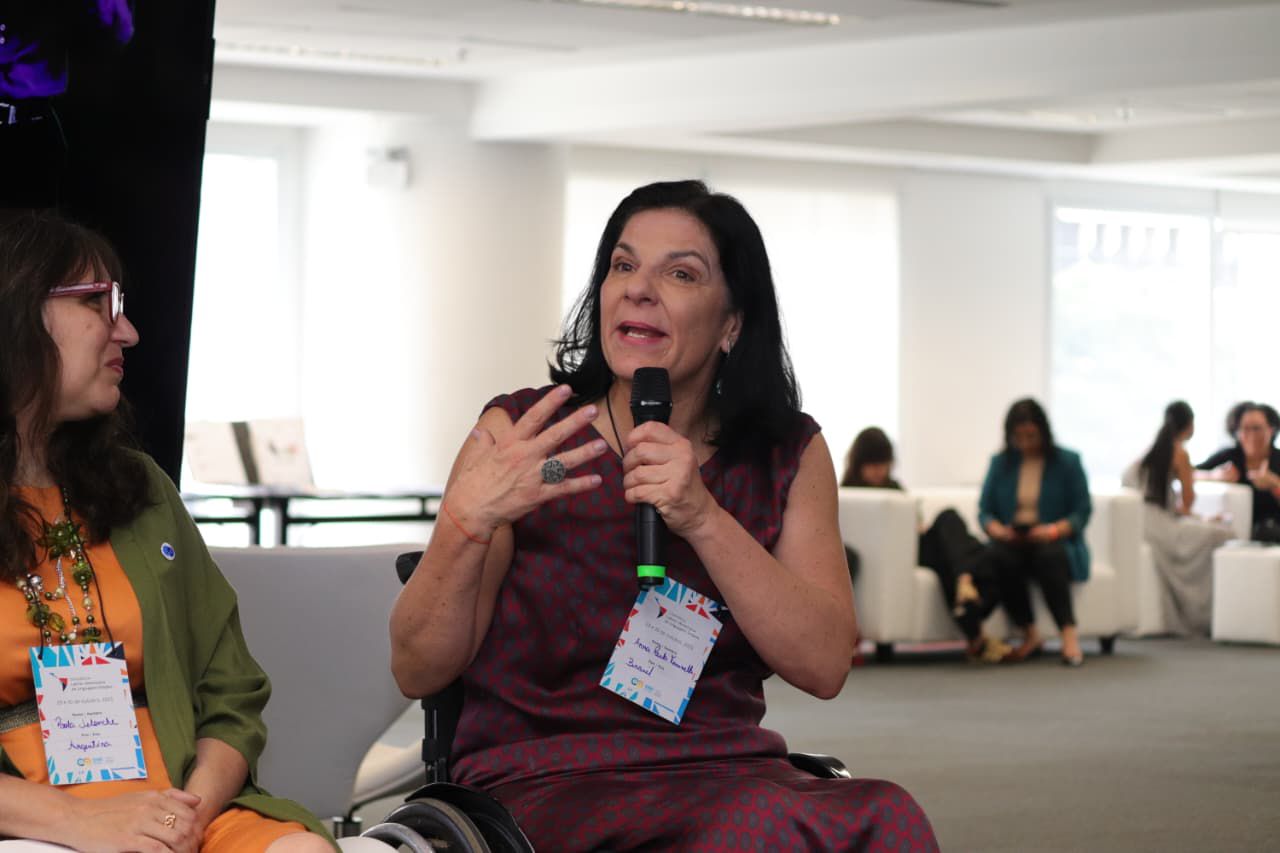 Anna Paula Feminella, secretária, está à direita, de vestido vinho, falando ao microfone no evento.