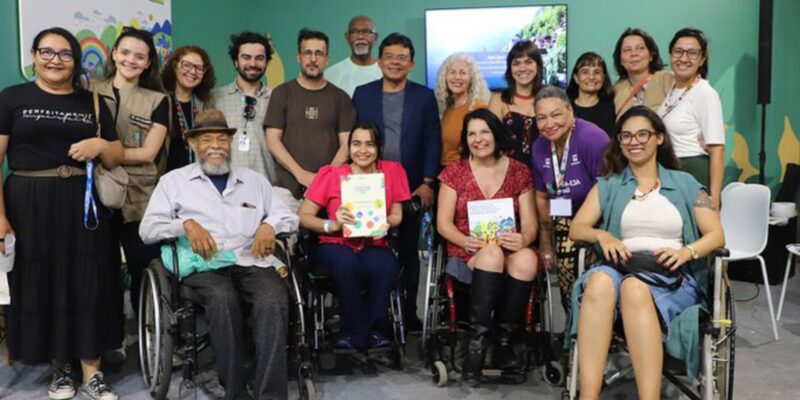 Grupo diverso, com pessoas em cadeiras de rodas, sorrindo em evento sobre direitos humanos na COP30.