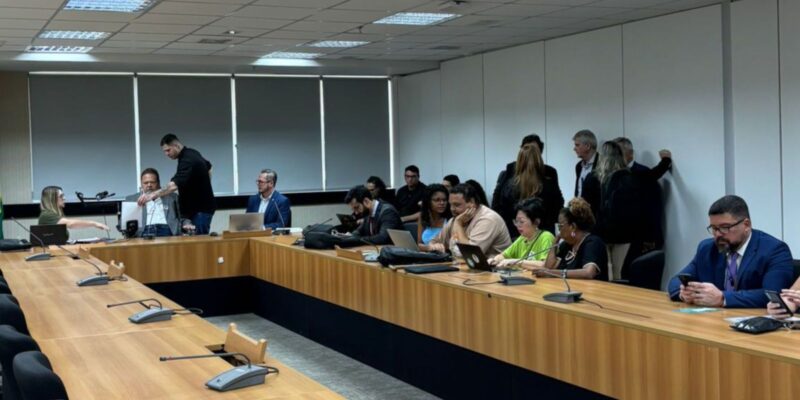 Sala de reunião, várias pessoas sentadas e algumas em pé, usando laptops e celulare