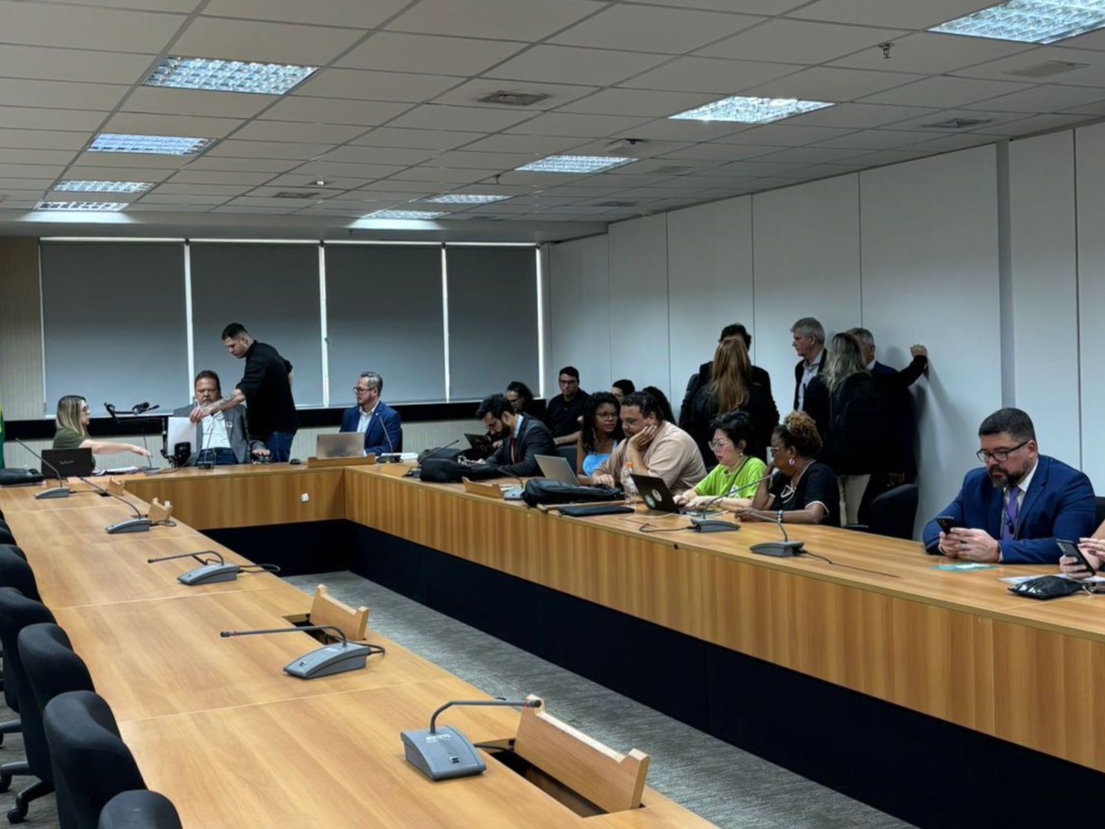 Sala de reunião, várias pessoas sentadas e algumas em pé, usando laptops e celulare