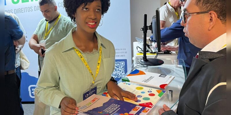 Isadora, mulher negra, sorri enquanto entrega revistas “Sem Limite” para visitante em estande de evento.