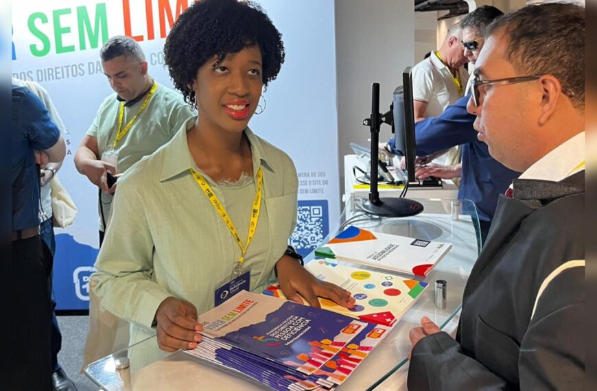 Isadora, mulher negra, sorri enquanto entrega revistas “Sem Limite” para visitante em estande de evento.