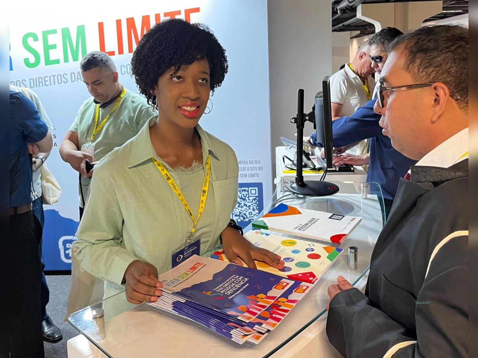Isadora, mulher negra, sorri enquanto entrega revistas “Sem Limite” para visitante em estande de evento.