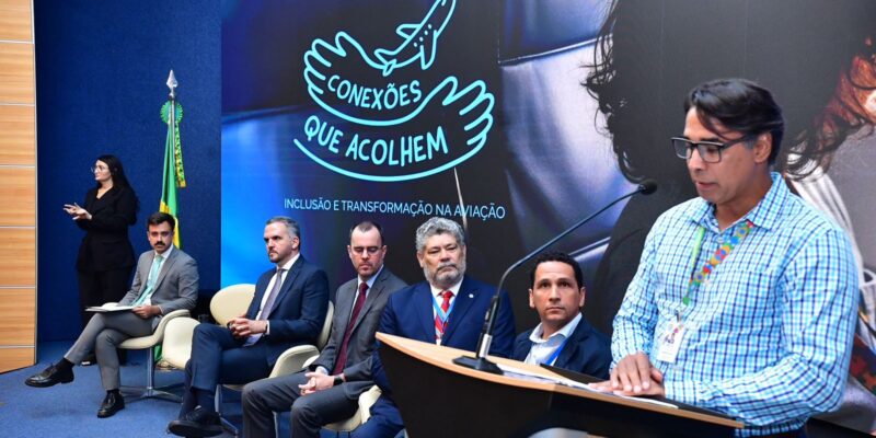 Evento formal com painel “Conexões que acolhem”, homens de terno sentados e um homem discursando.