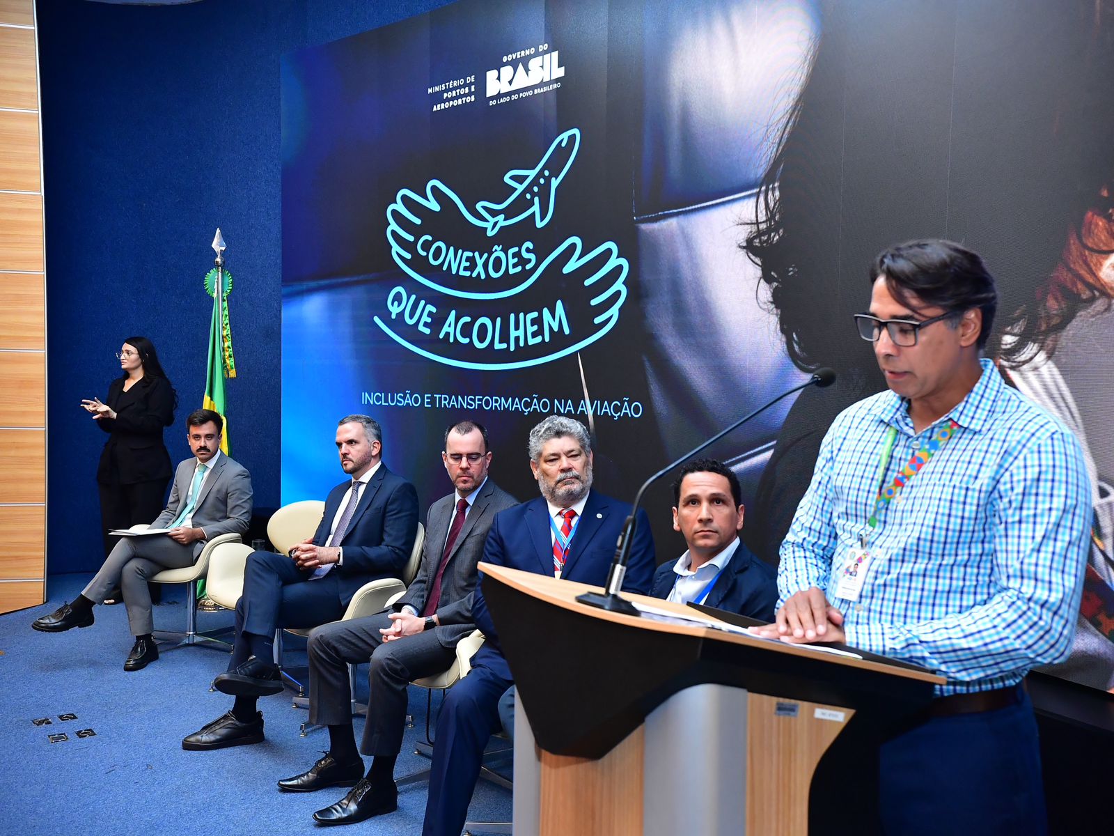 Evento formal com painel “Conexões que acolhem”, homens de terno sentados e um homem discursando.