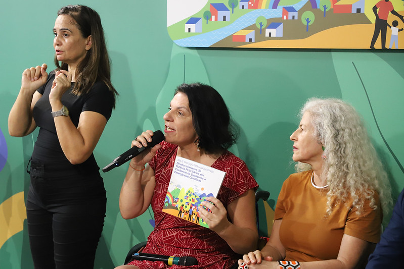 Ministério promoveu roda de conversa, visitas técnicas e articulações estratégicas durante o evento, com foco em acessibilidade, gestão de risco e tecnologias assistivas Três mulheres em evento; uma faz Libras, outra fala ao microfone e segura um livreto colorido.