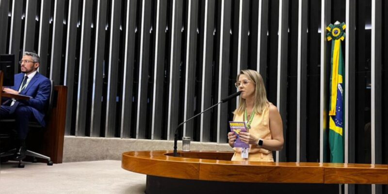 Mulher discursa em tribuna legislativa com bandeira do Brasil ao fundo; homem trabalha ao lado.