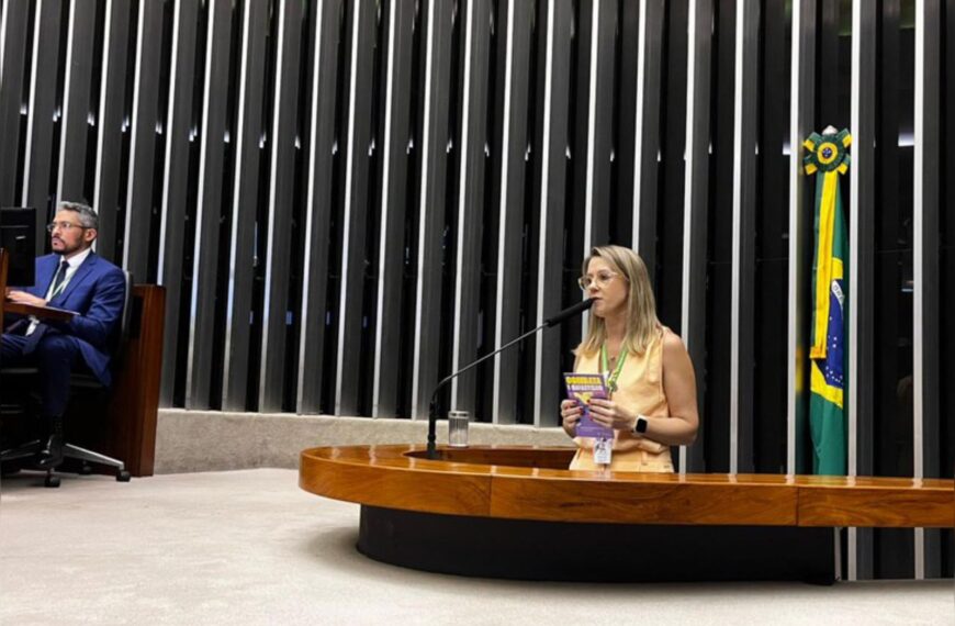 Mulher discursa em tribuna legislativa com bandeira do Brasil ao fundo; homem trabalha ao lado.