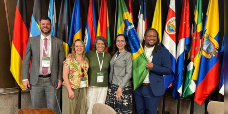 Cinco pessoas sorrindo em pé, com crachás, diante de várias bandeiras de países.