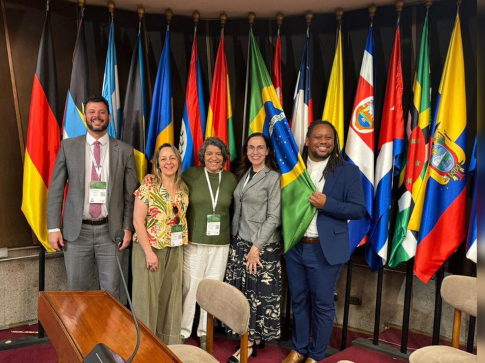 Cinco pessoas sorrindo em pé, com crachás, diante de várias bandeiras de países.