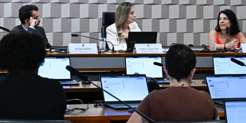 Reunião formal com três pessoas na mesa principal e duas de costas, usando notebooks.