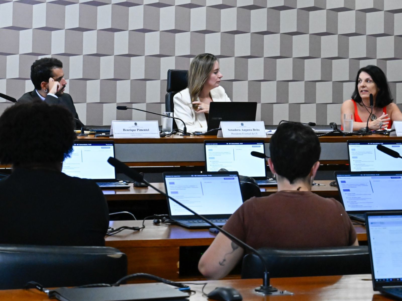 Reunião formal com três pessoas na mesa principal e duas de costas, usando notebooks.