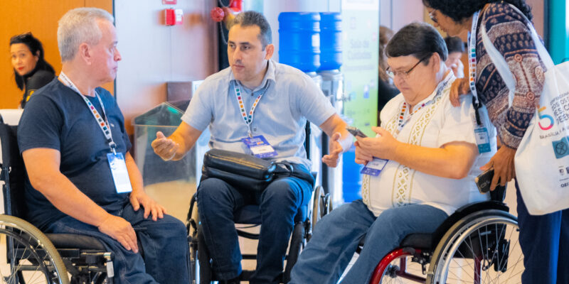 Três homens em cadeiras de rodas conversam animados; um deles mexe no celular. Uma mulher, de pé, observa. Todos usam crachá, sugerindo participação em evento inclusivo e acessível.