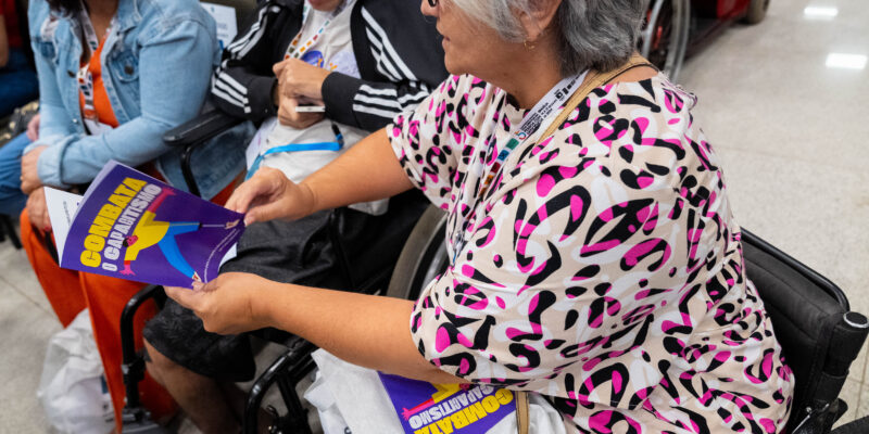 Senhora de cabelos grisalhos e blusa colorida está em uma cadeira de rodas, lendo um livreto com capa roxa e título “Combata o Capitalismo”. Outras pessoas e mais exemplares estão ao redor.