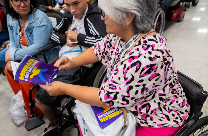 Senhora de cabelos grisalhos e blusa colorida está em uma cadeira de rodas, lendo um livreto com capa roxa e título “Combata o Capitalismo”. Outras pessoas e mais exemplares estão ao redor.