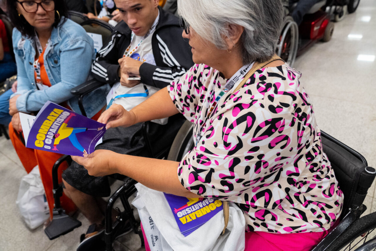Senhora de cabelos grisalhos e blusa colorida está em uma cadeira de rodas, lendo um livreto com capa roxa e título “Combata o Capitalismo”. Outras pessoas e mais exemplares estão ao redor.