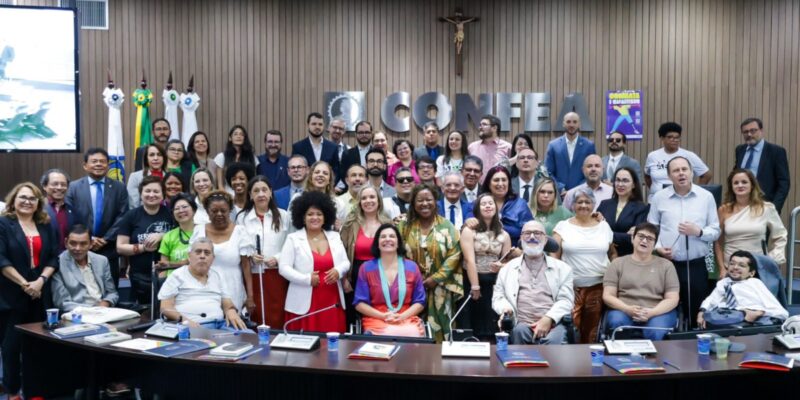 Foto com grande grupo diverso em auditório, incluindo pessoas com deficiência. Letreiro “CONFEA” ao fundo, clima formal, materiais sobre a mesa e atmosfera de evento inclusivo.