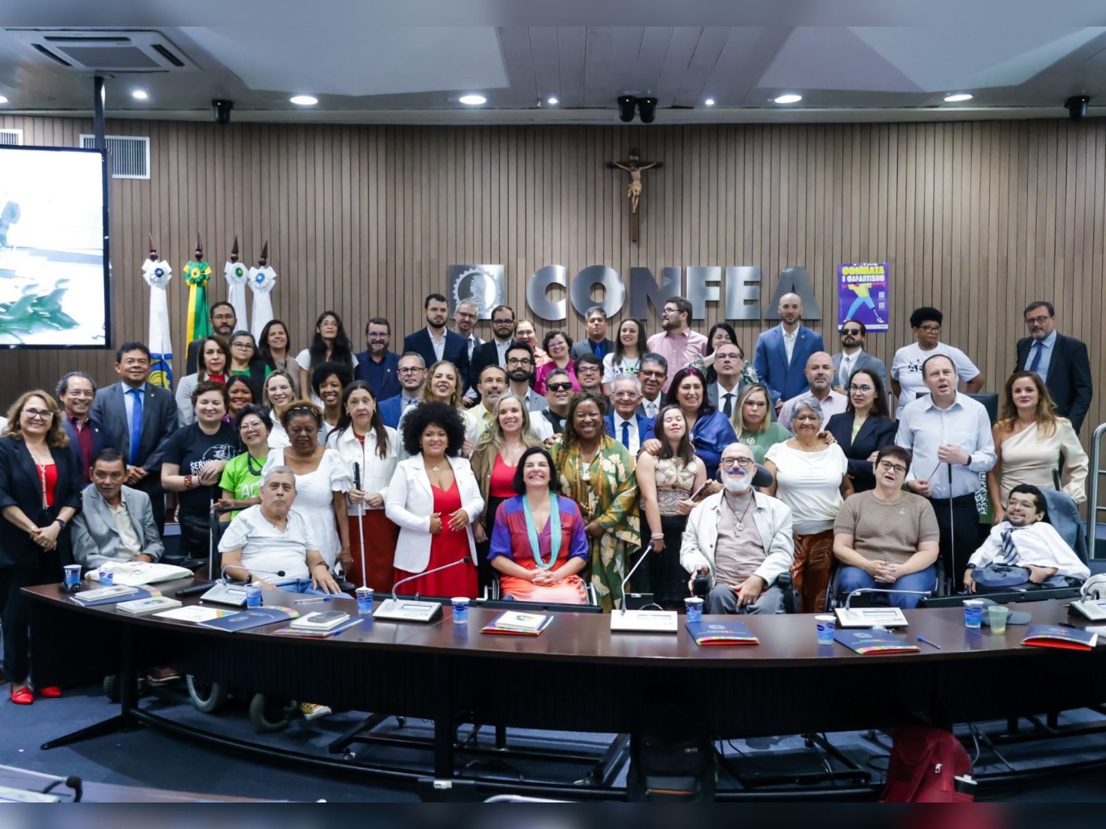 Foto com grande grupo diverso em auditório, incluindo pessoas com deficiência. Letreiro “CONFEA” ao fundo, clima formal, materiais sobre a mesa e atmosfera de evento inclusivo.