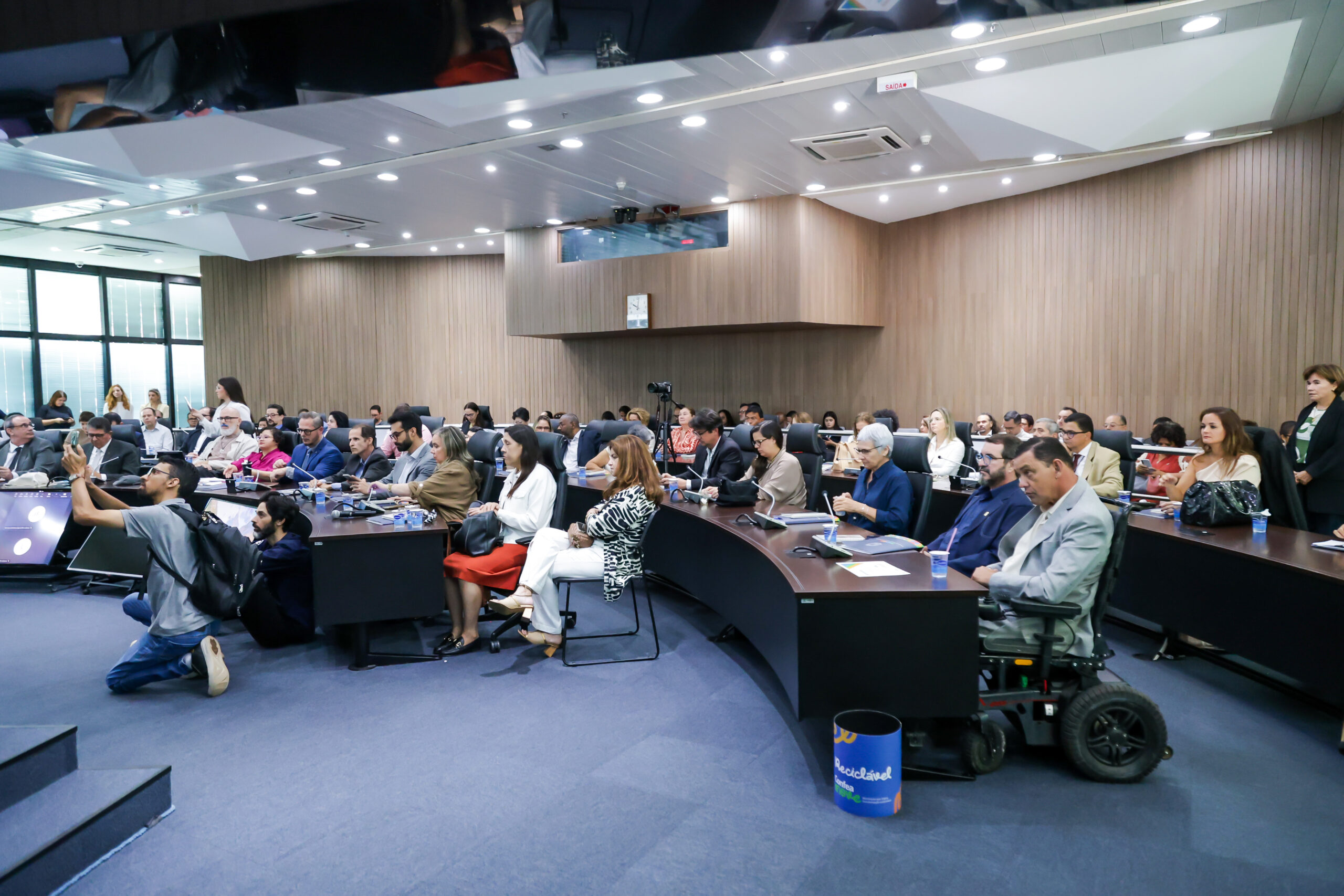 Conade empossa novos conselheiros e reforça agenda de inclusão Sala de reunião cheia, pessoas em traje social sentadas em mesas em U. Ambiente moderno, bem iluminado. Homem em cadeira de rodas motorizada e fotógrafo à esquerda.
